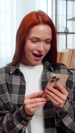 young woman sitting on sofa scrolling news feed on smartphone occasionally smiling reading updates
