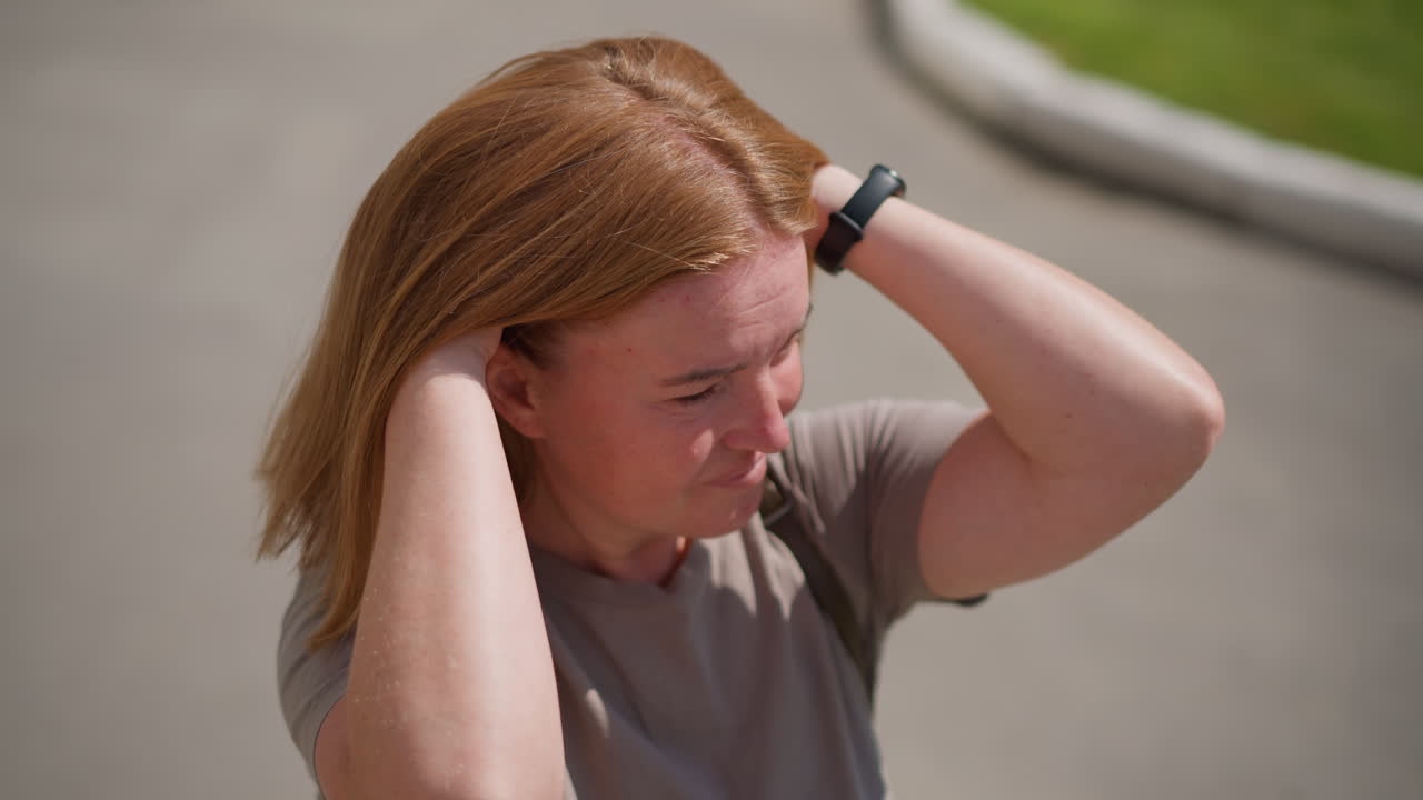 Top view of lady scratching hair with both hands wearing smart watch under sunlight near sidewalk and green grass, subtle body language expresses discomfort, reflection, and emotional stress