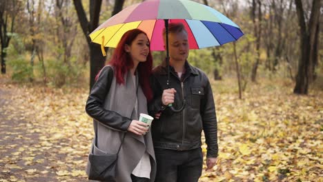 young happy couple walking together in autumn park holding a colorful umbrella. attractive woman with red hair is holding a paper cup with coffee. they are talking and holding hands