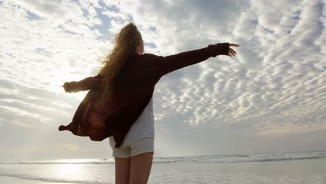 Rear-view-of-young-woman-with-arms-outstretched-looking-at-sea-on-the-beach-4k