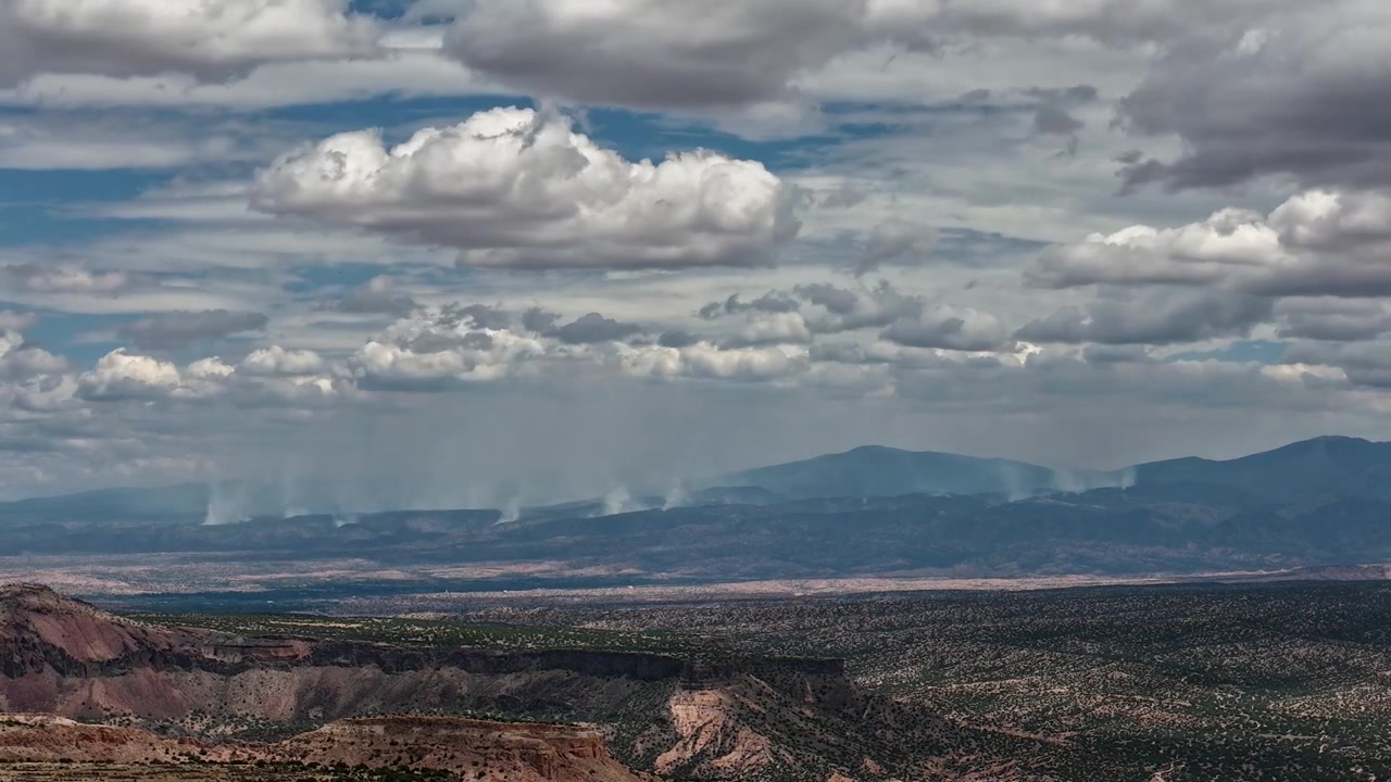 Timelapse 4K con drone che cattura molteplici pennacchi di fumo che si innalzano dall'incendio di Frijoles nelle montagne distanti. La vista panoramica mostra scogliere a mesa e canyon profondi sotto una fitta coltre di nuvole