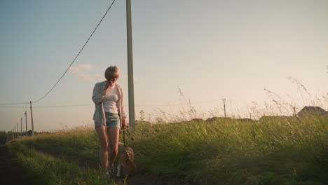 femme en short de jean marchant sur un appel téléphonique tôt le matin à travers un champ herbeux, tenant une laisse dans une main, avec en arrière-plan une verdure, un poteau électrique et un bâtiment rural lointain