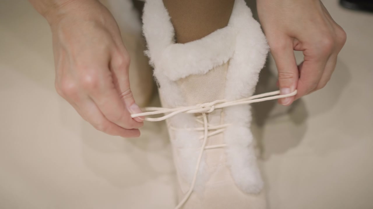 Close up of hands tying laces on white fur lined boot. person manipulates cream colored shoe laces creating and tightening them around ankle high footwear. Soft lighting highlights details