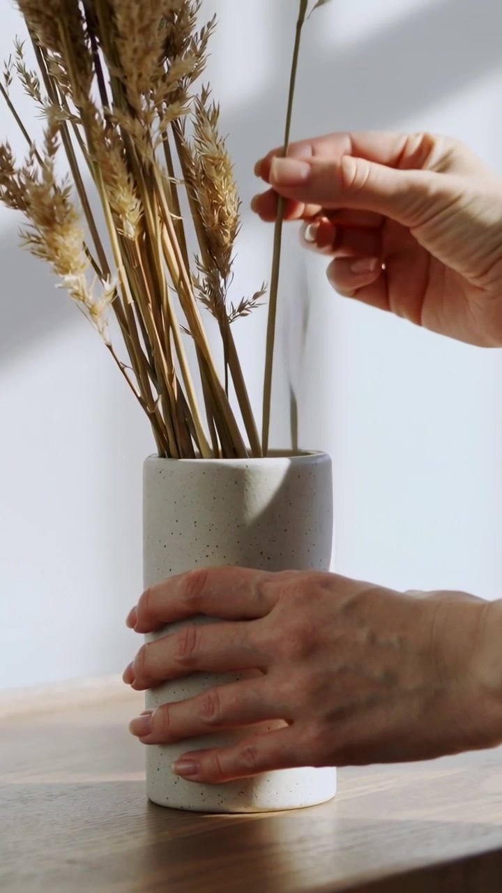 Vidéo verticale : Mains arrangeant des tiges séchées dans un vase blanc sur une table à la maison pour la décoration.