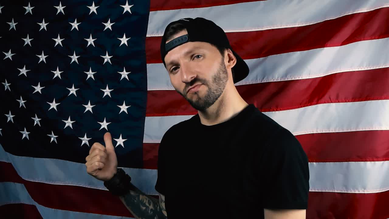 Proud american patriot with tattoos and short beard points slowly at an American United States flag that is behind him slowly moving in the wind. Man is nodding with approval, wearing black clothing