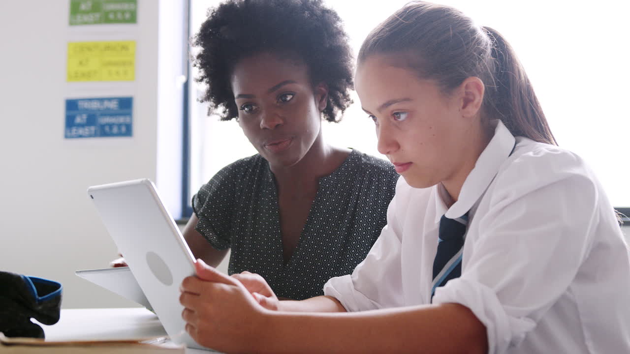 tutora de escuela secundaria con tableta digital dando a una estudiante que lleva uniforme una a una enseñanza en el escritorio