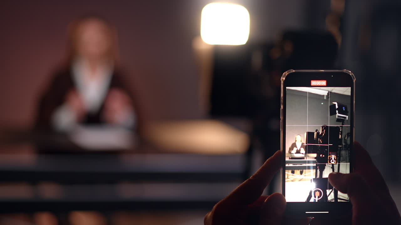Footage on smartphone in photo studio. Close up. Woman sits at desk in blurred backdrop. Blogging concept.