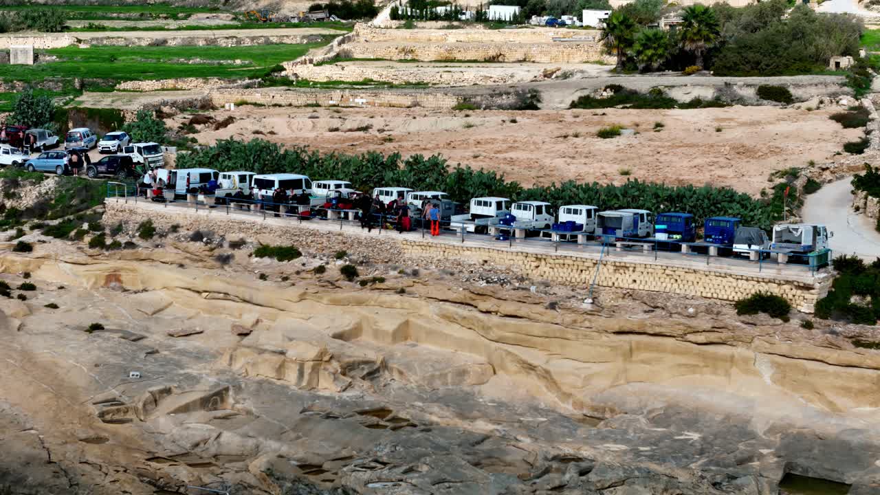 Una fila di furgoni e camioncini bianchi e blu parcheggiati si trova in cima alle erose scogliere calcaree di Gozo, Malta, con vista su una valle costiera rocciosa sotto il brillante sole estivo del Mediterraneo