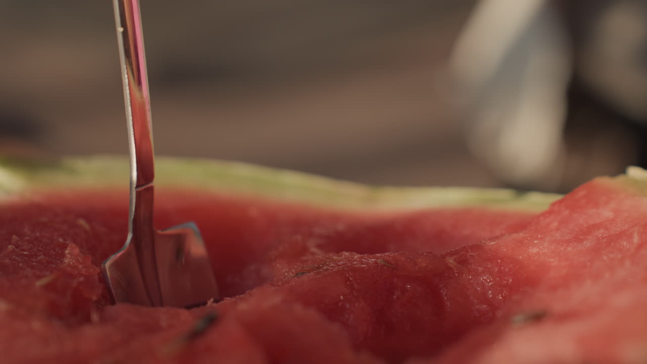 Primo piano di una chef bianca che raccoglie un pezzo di anguria con una paletta di metallo, polpa rossa compatta compressa e poi sollevata in un cucchiaio luccicante, l'illuminazione del patio della cucina mette in risalto la texture, attenzione culinaria sulla porzione