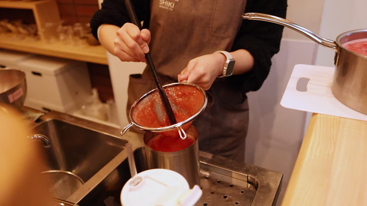 Um chef japonês com um avental marrom coa purê de frutas vermelhas através de uma peneira de metal.