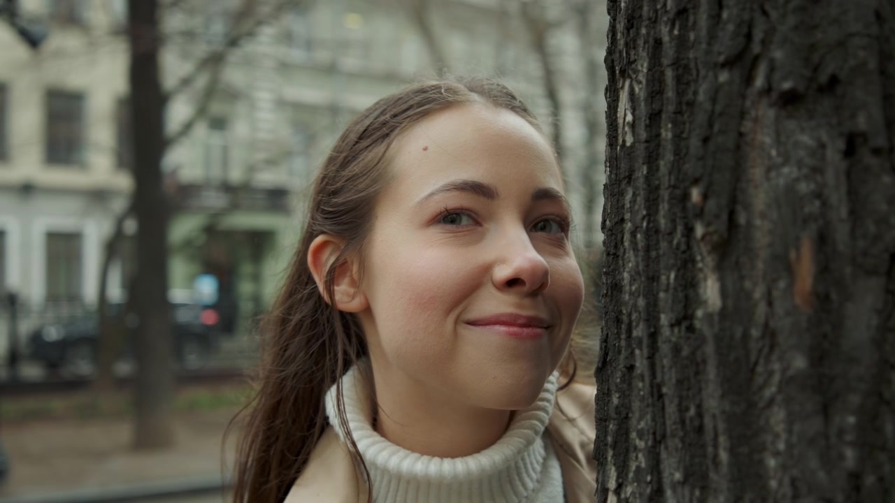Primo piano di una giovane donna con lunghi capelli castani che indossa un dolcevita bianco e un cappotto beige, in piedi all'aperto vicino a un albero. La scena presenta una luce naturale soffusa, edifici urbani sullo sfondo e minimale.