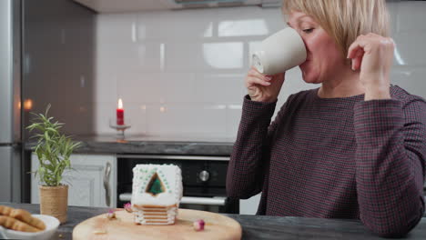 mujer bebe de la taza mientras descansa con la cabeza en la mano, mirando pensativamente a la casa de pan de jengibre en una bandeja de madera, luz de vela cálida, planta en olla y entorno de cocina rústica