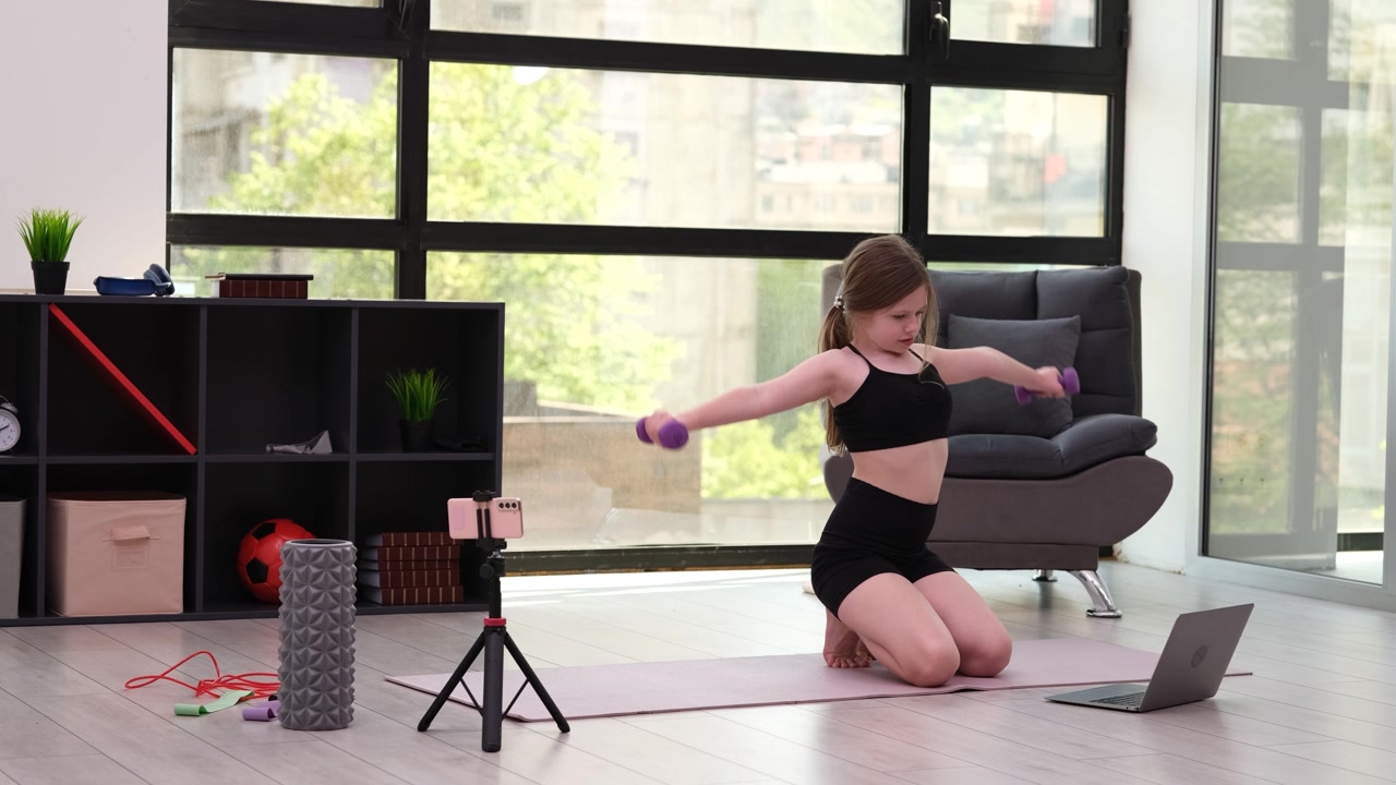 Girl sits on yoga mat and follows movements on screen of laptop