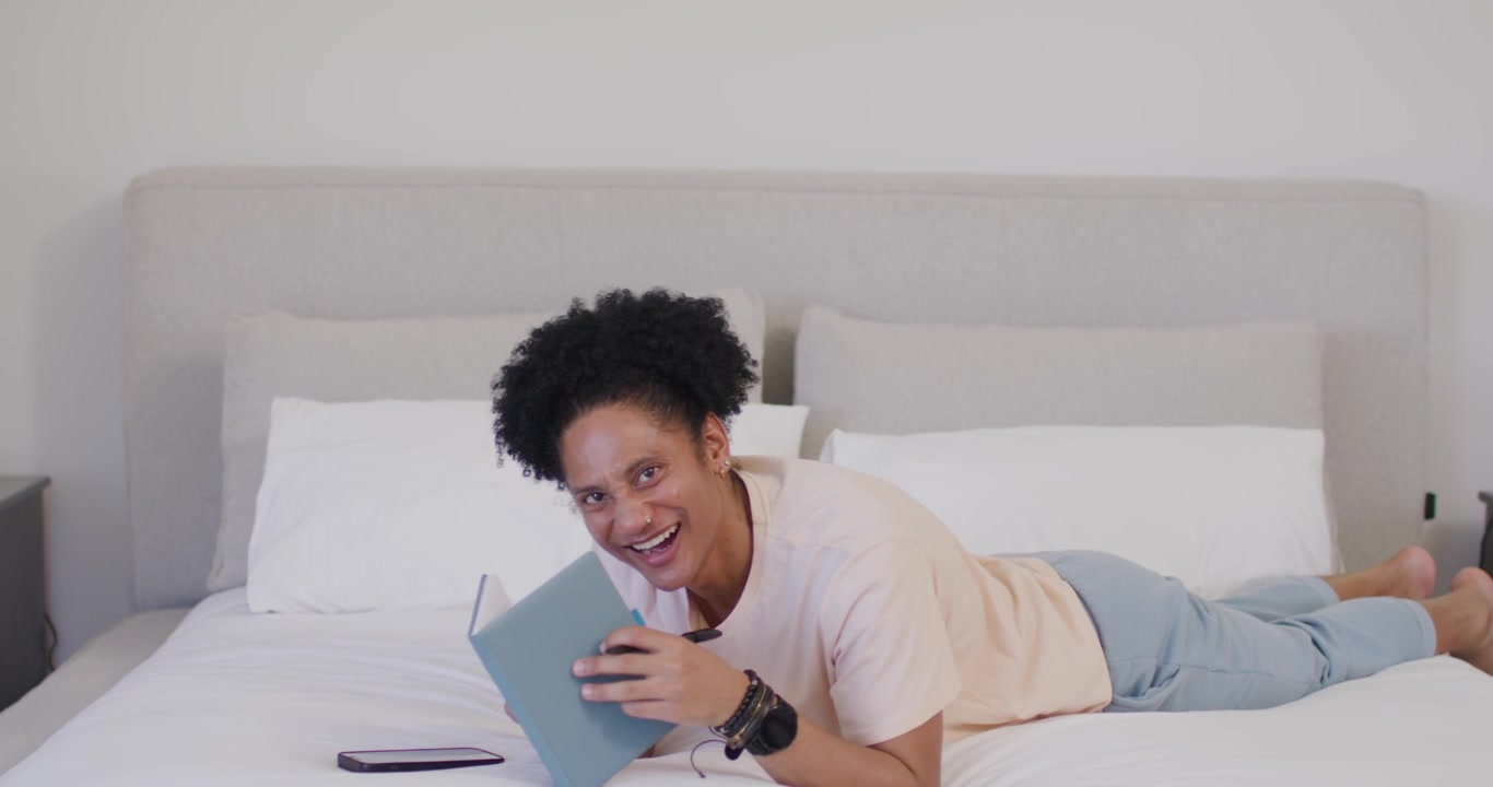 African American adult male lying on bed writing in blue notebook, glancing at smartphone, smiling