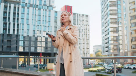 happy mature businesswoman in wireless headphones dancing listening to music on downtown street