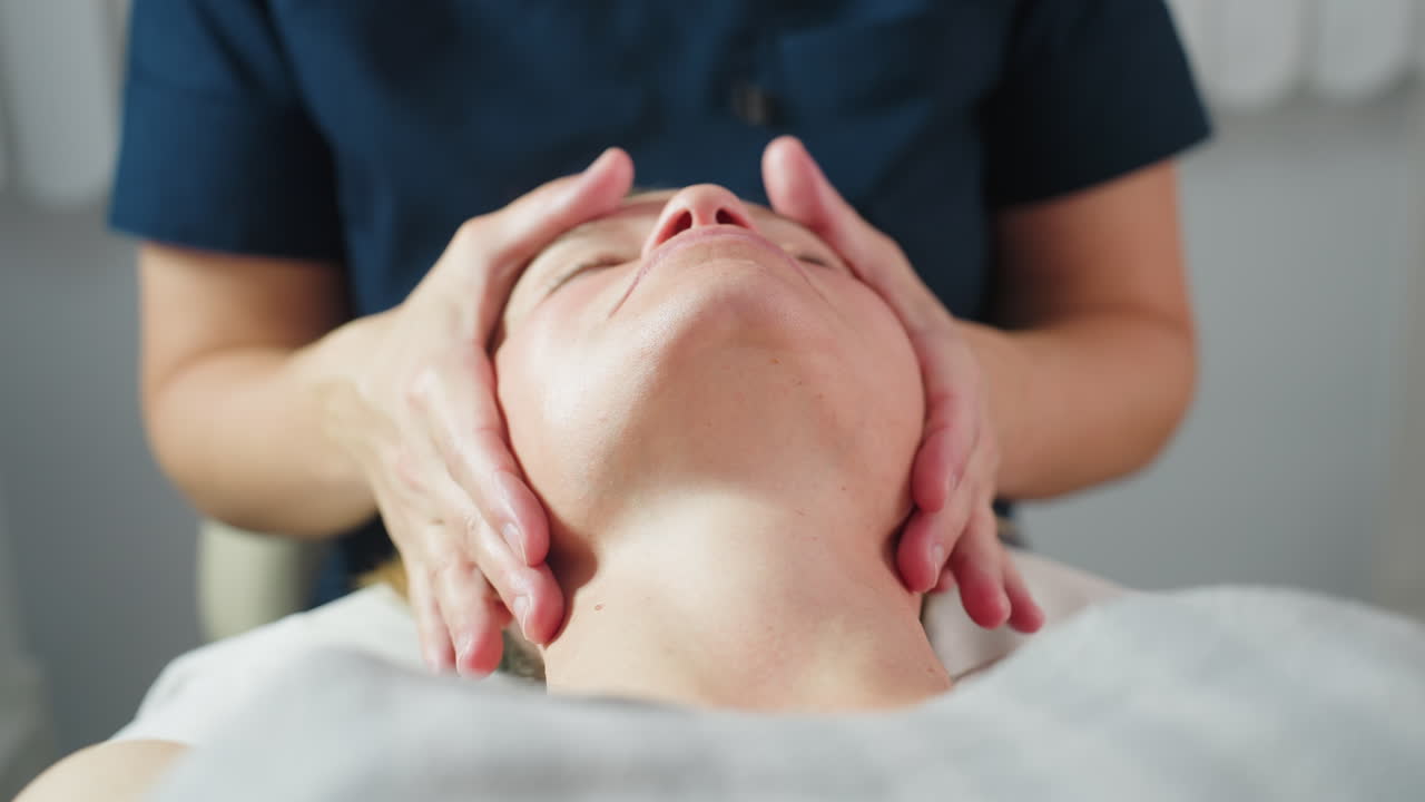 Professional tilts client head gently in bright spa room with soothing white background during relaxing facial massage therapy session showcasing expert hands and tranquil wellness environment