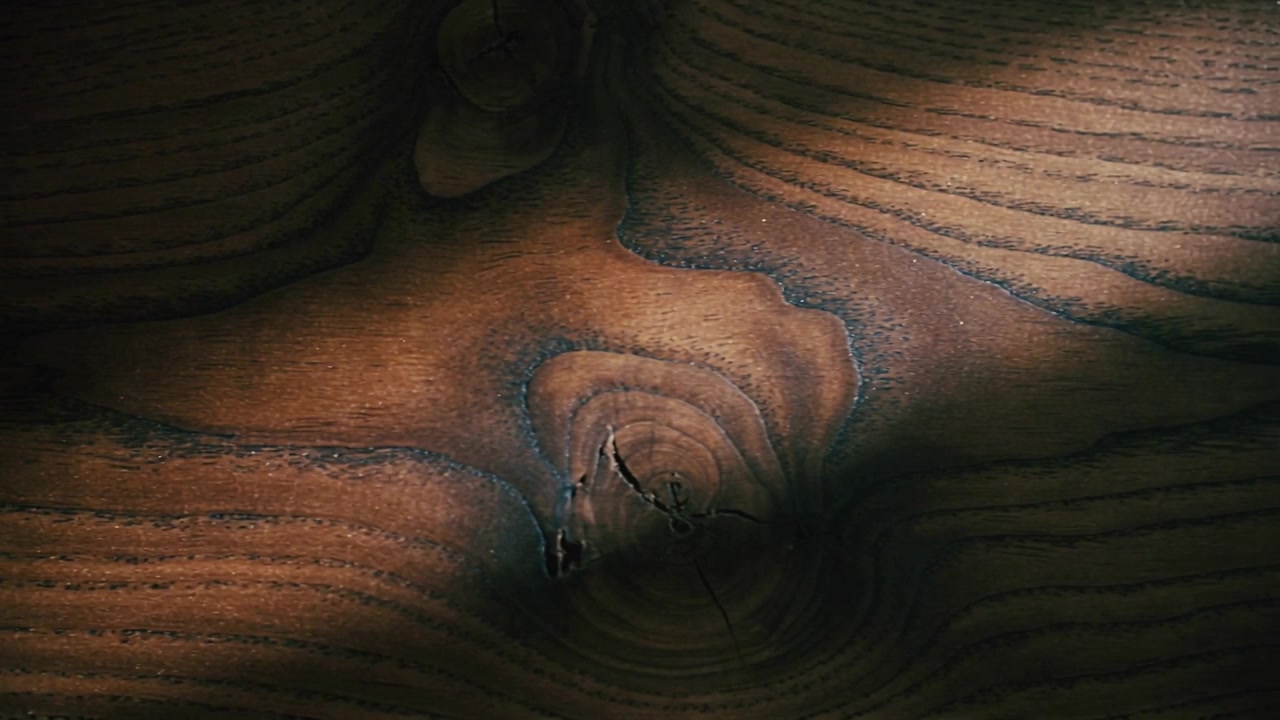 Luce cangiante che rivela un primo piano di un'asse di legno macchiata in un interno, mostrando un nodo centrale con crepe.