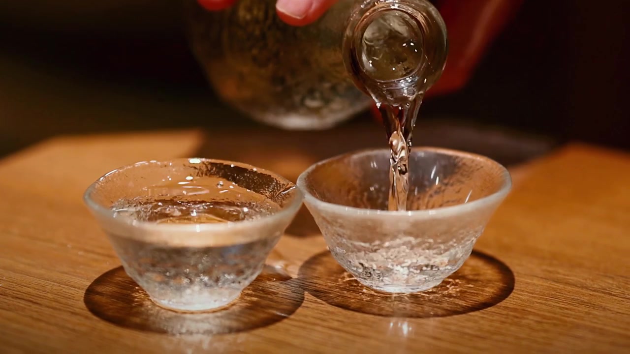 Seseorang menuangkan arak beras bening dari teko kaca ke dalam dua cangkir kecil bertekstur