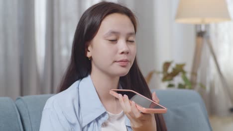 close up of asian teen girl recording voice message on smartphone while sitting on sofa in the living room at home