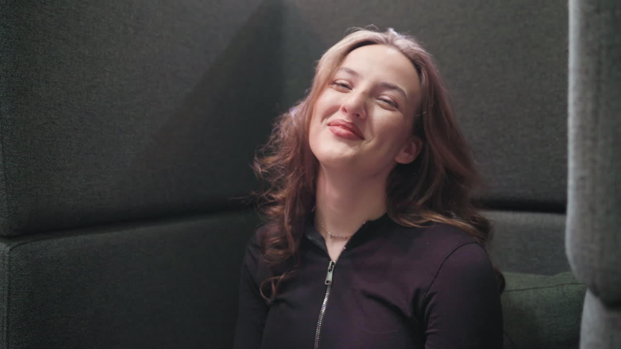 Light skin lady with blond hair smiles brightly while looking directly at camera, seated comfortably against dark cushioned wall in relaxed indoor setting, exuding playful and cheerful expression