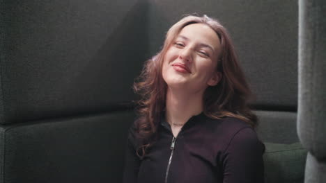 light skin lady with blond hair smiles brightly while looking directly at camera, seated comfortably against dark cushioned wall in relaxed indoor setting, exuding playful and cheerful expression