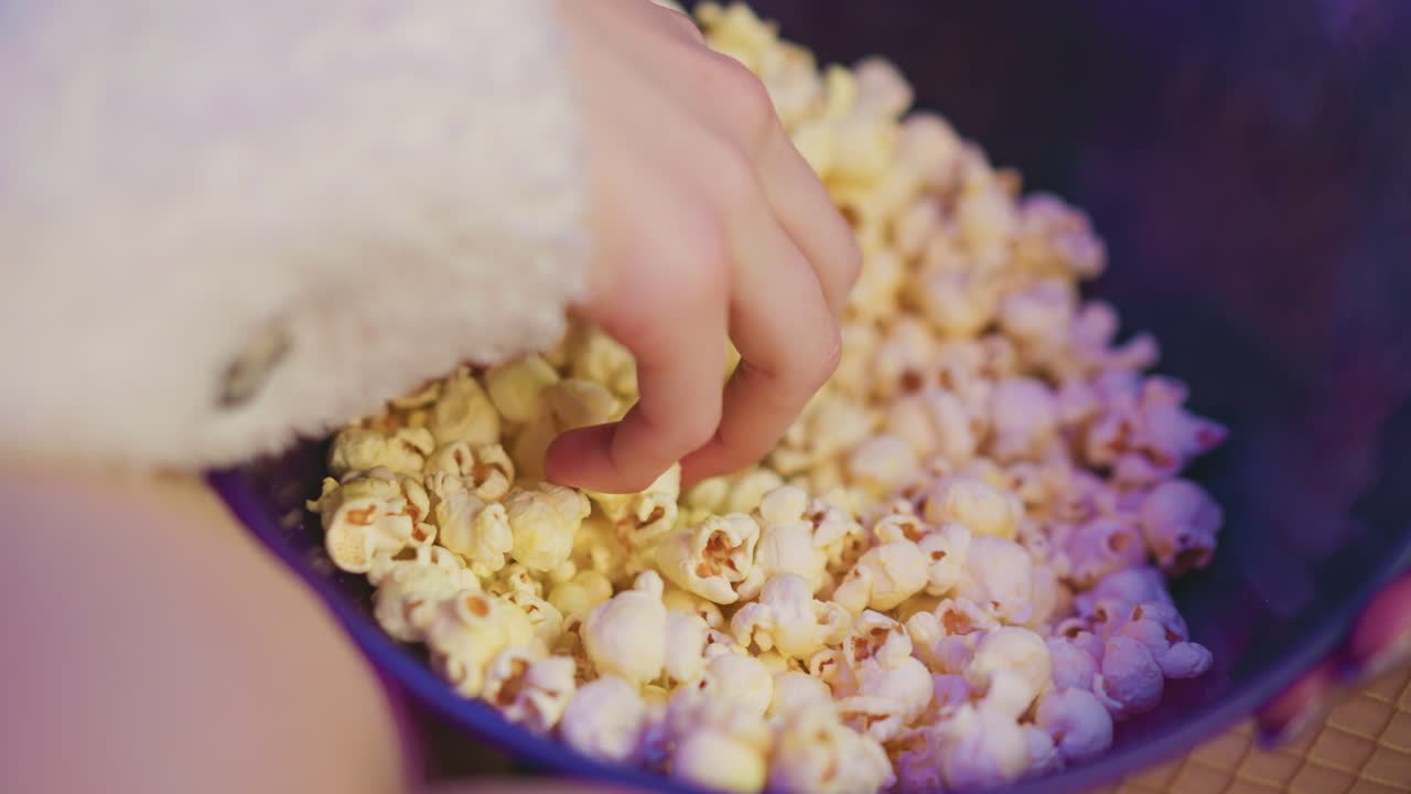 Close up of fresh popcorn in transparent bowl under soft purple lighting as someone reaches into it to grab snack, creating cozy relaxed ambiance in dim indoor setting