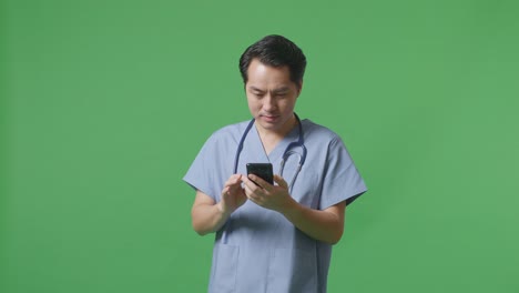 병원 에서 녹색 화면 배경 에 서 있는 동안 스마트폰 을 사용 하는 스테토스코프 를 들고 있는 아시아인 남성 의사