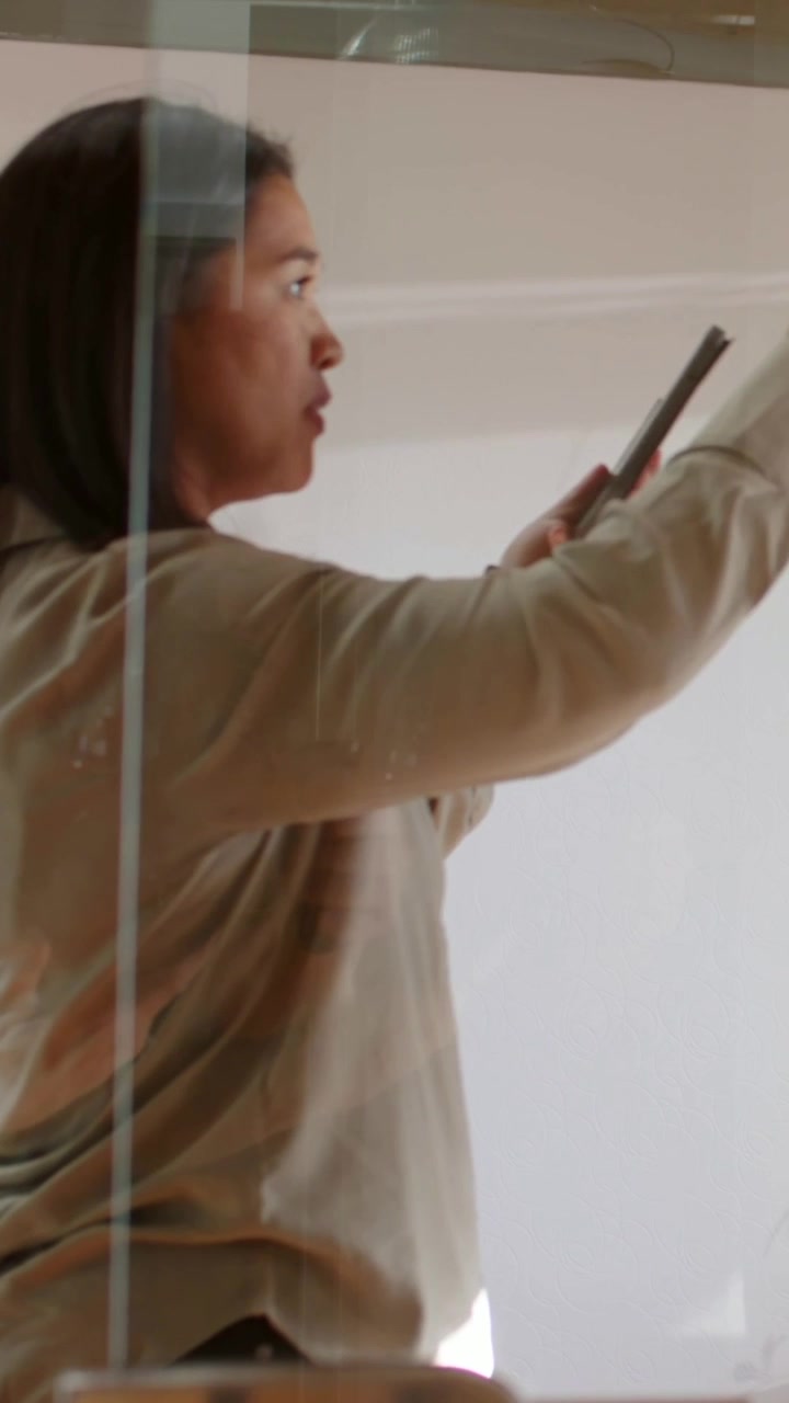 Video verticale: Donna che indica dopo aver dato un'occhiata a un tablet, gesticolando in una sala riunioni, con un top color cuoio, dietro un vetro.