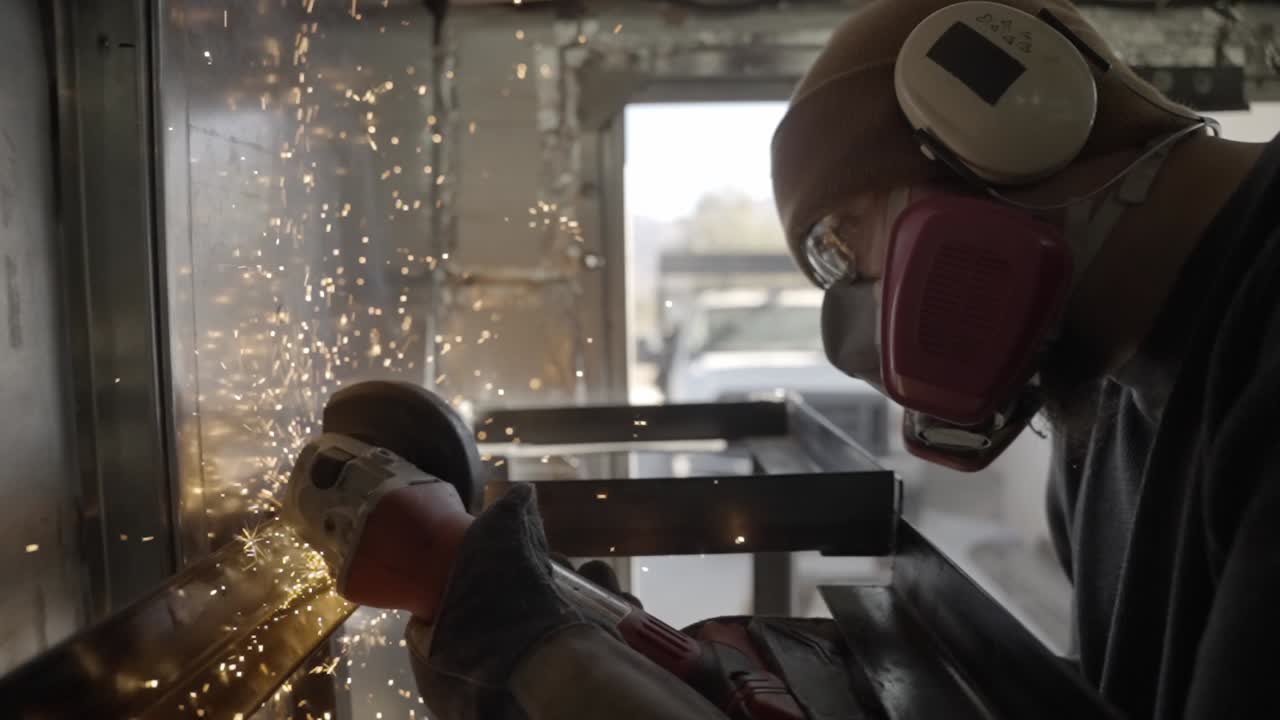 Worker wearing protective gear grinds metal shelves with power tool, sparks fly- slow mo