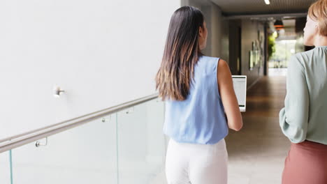 walking down hallway, two women discussing work while holding laptop