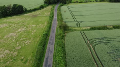 A-slow-descending-and-camera-rising-shot-revealing-a-car-speeding-down-a-narrow-country-lane-between-two-large-fields-on-an-overcast-day-in-england