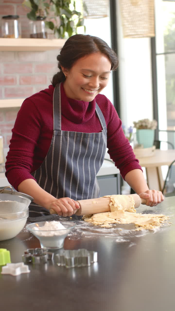 Vertical video of happy biracial woman in apron rolling dough in kitchen at home, slow motion