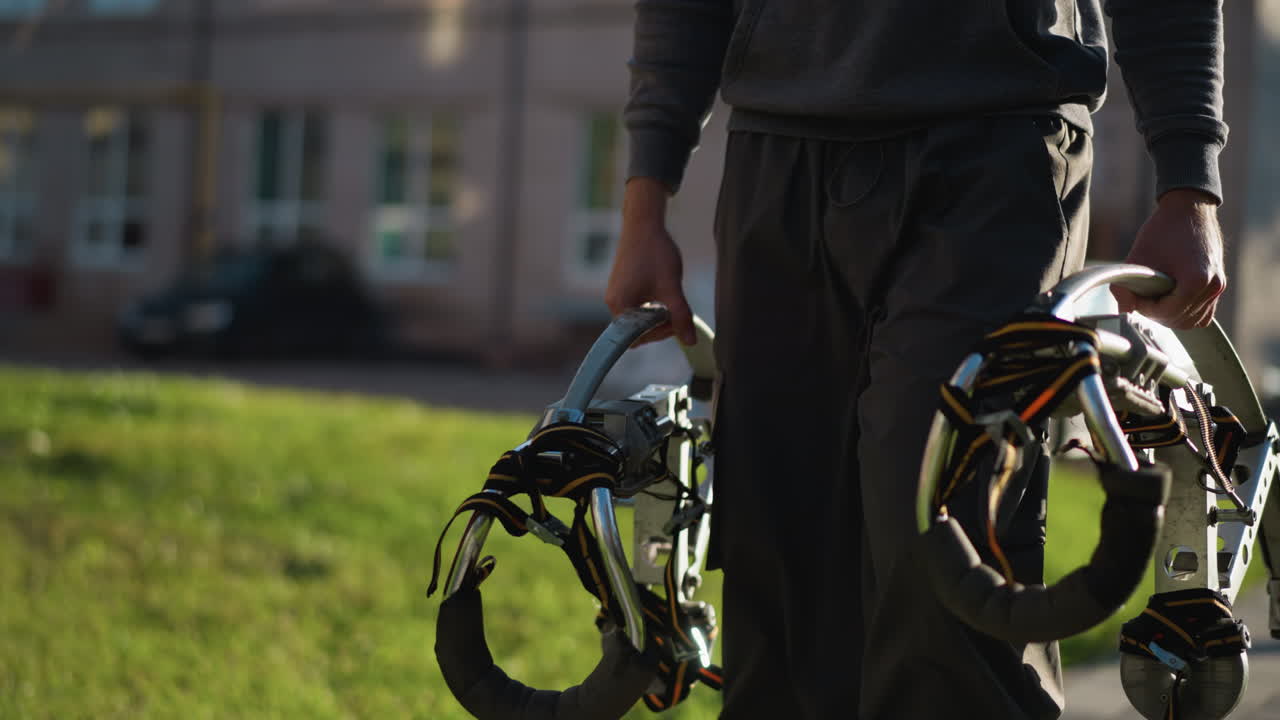 어두운 회색 스웨트 팬츠와 긴 팔 셔츠를 입은 남자가 큰 금속 기둥을 들고, 뒷면에 흐릿한 녹색 풀과 주거 건물과 함께 야외에 서 있습니다.