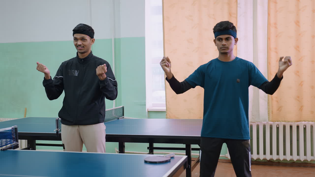 Dua pemain tenis muda dengan pakaian olahraga melakukan latihan pemanasan tangan sebelum pertandingan, berdiri di samping meja dengan raket yang diletakkan di atas permukaan