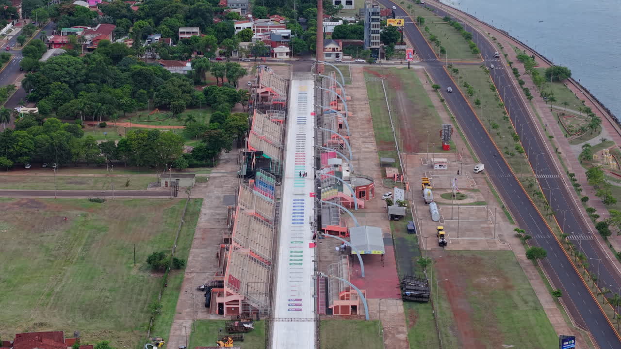 Wide angle daisy cutter drone clip over Sambodromo in Encarnación, Paraguay. Scene shows full stage length, with road and river on right and vegetation on left.