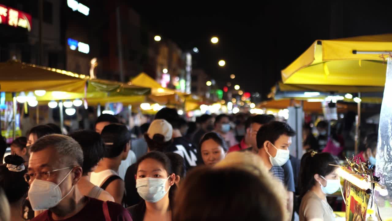 A bustling night market in Kuala Lumpur with masked people walking through food stalls, exploring local cuisine.