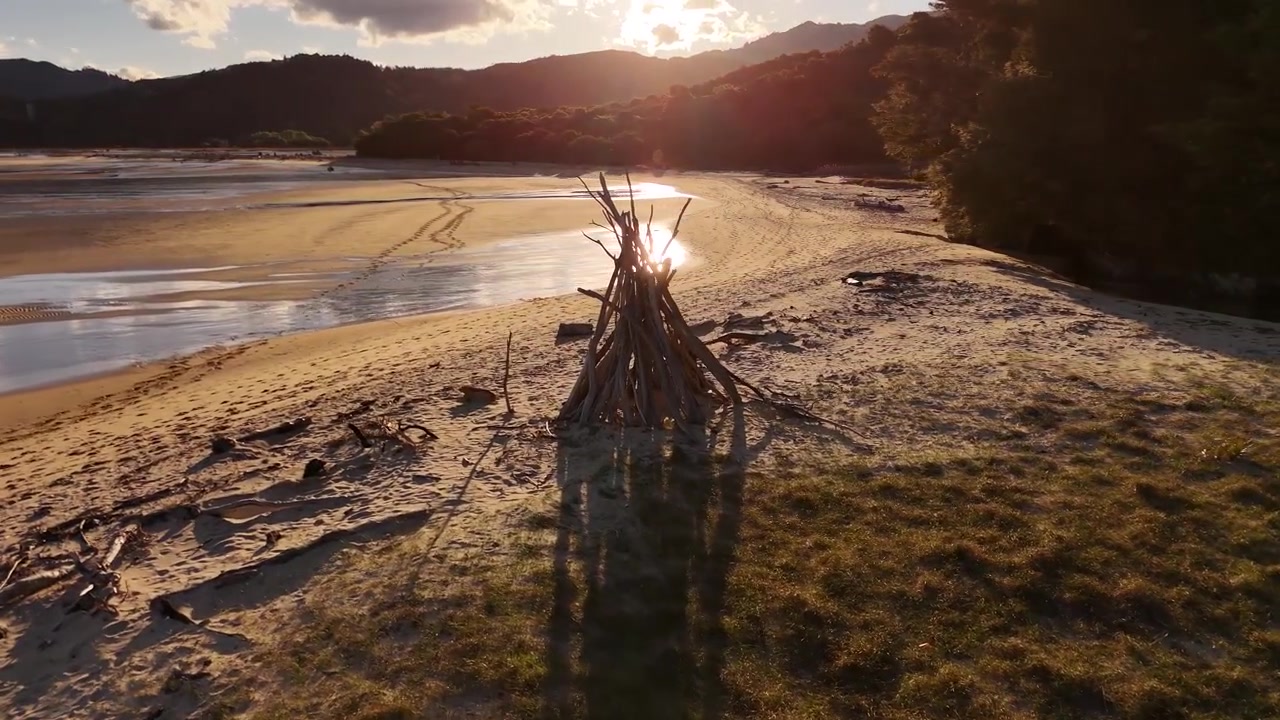 Treibholz-Feuerstelle auf einer weiten Gezeiten-Sandfläche mit flachen spiegelnden Wasserbecken, einer Flussmündung und buschbewachsenen Hügeln, die im goldenen Sonnenuntergangslicht im Abel Tasman Nationalpark, Neuseeland, leuchten. Luftaufnahme.