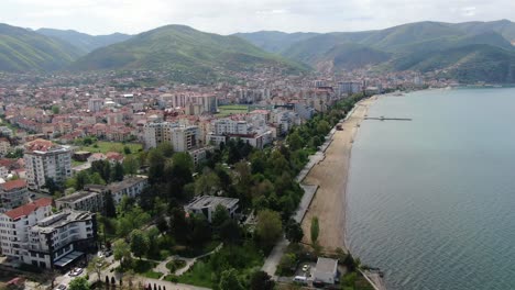 drone view in albania flying over ohrid lake in pogradec beach town with apartment buildings, green mountains and sandy shore