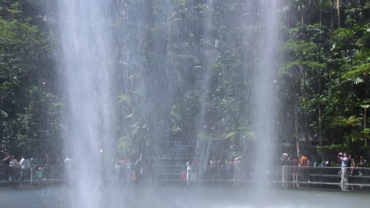 People stand on a viewing platform watching a large indoor waterfall cascade in a bright, verdant garden environment with natural daylight and steady camera