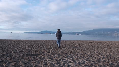 youg woman wearing black beanie, black puffy coat and jeans on a beach walking towards the sea on a beautiful winter day with boats and oil tankers in the background