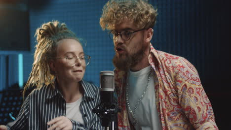 portrait shot of the stylish young man and woman in hipster style, singers of the duo singing in the microphone and recording a song in the sound studio