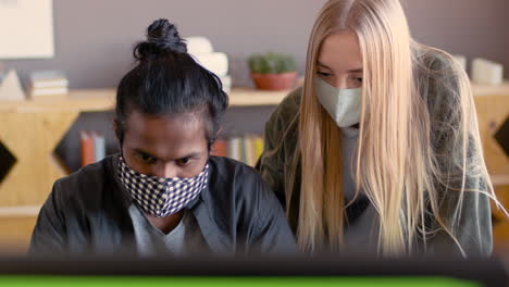 two multiethnic colleagues with face masks looking at computer screen and discussing a new project in the office