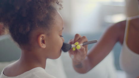 close up of a little african american girl having her mum doing make-up on her, applying powder on her cheeks with makeup brush