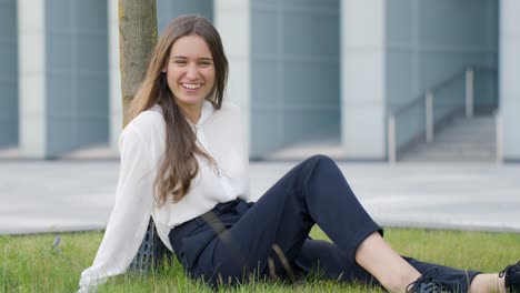 Frau-Macht-Alberne-Gesichter,-Während-Sie-Im-Gras-Sitzt-Und-Sich-An-Einen-Baum-Dahinter-Lehnt,-Statische-Aufladung