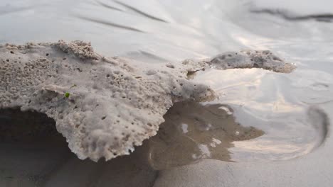 close up footage of water flowing and bubbling out from sandy beach ground that has a chunk or a piece of ice with sand covering it on top. water is reflective and bubbling and flowing in slow motion.