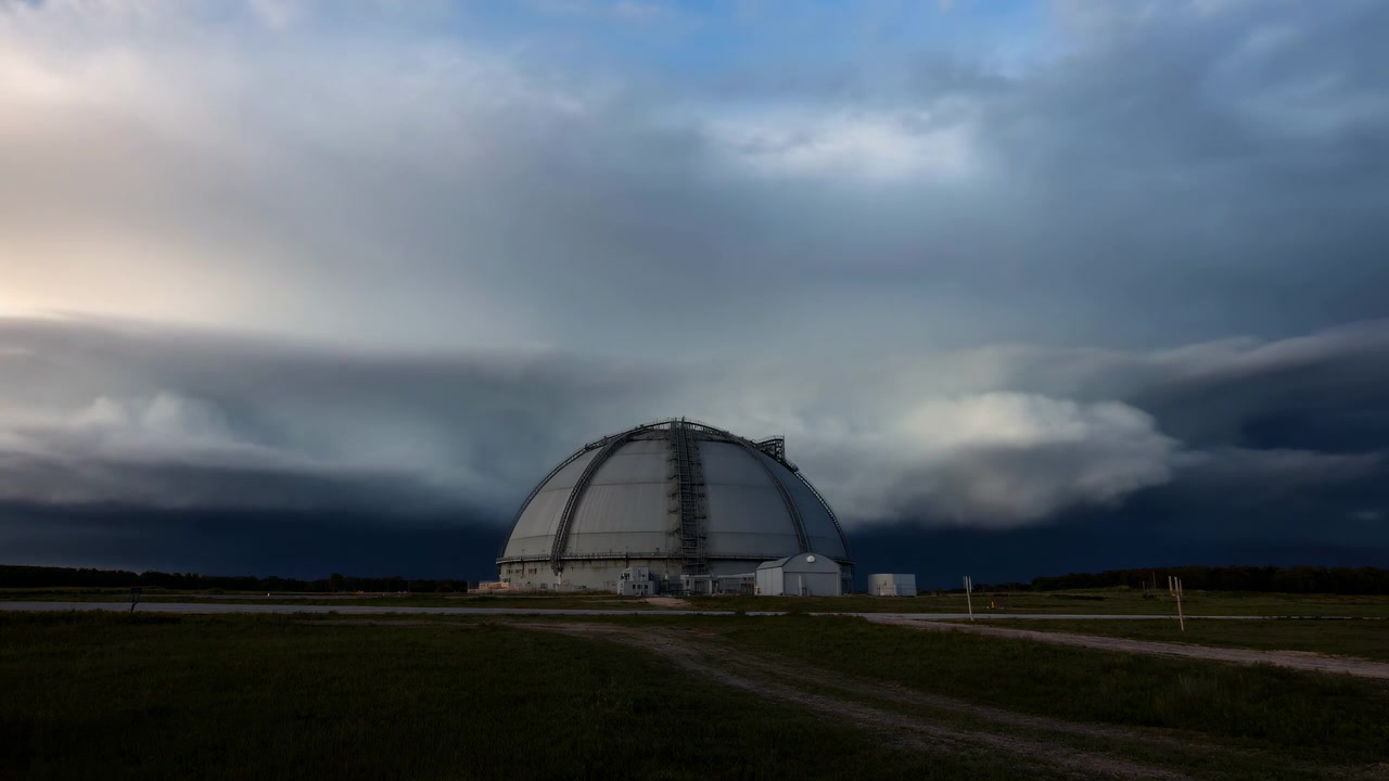 En stor hvid industridome står på en ryddet serviceplads på en flad græsmark, mens lagdelte stormskyer skifter fra lyse solbelyste højdepunkter til tiltagende mørke, hvilket synligt reducerer belysningen.