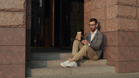 Relaxed-Businessman-Drinking-Coffee-And-Listening-Music-On-The-Smartphone-Via-Earphones-While-Sitting-On-Steps-Of-A-Building-In-The-City-1
