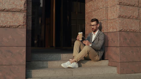 relaxed businessman drinking coffee and listening music on the smartphone via earphones while sitting on steps of a building in the city 1