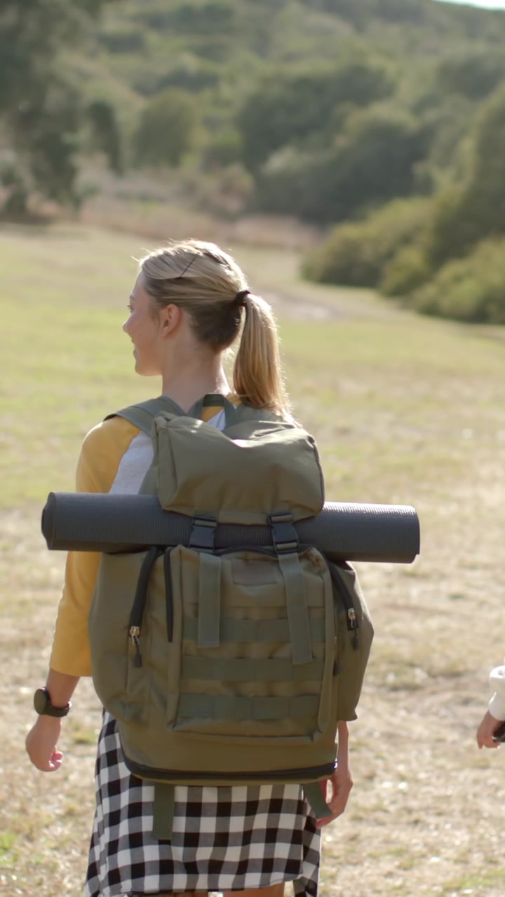 Vertical video: Female friends walking trail, lead turning, talking, carrying olive pack and mat