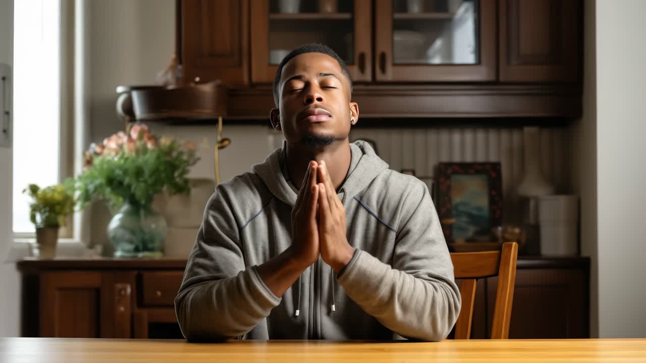 Hopeful young man sitting at a kitchen table, eyes closed and hands clasped, praying for peace and guidance while expressing deep gratitude and devotion in a serene moment of reflection